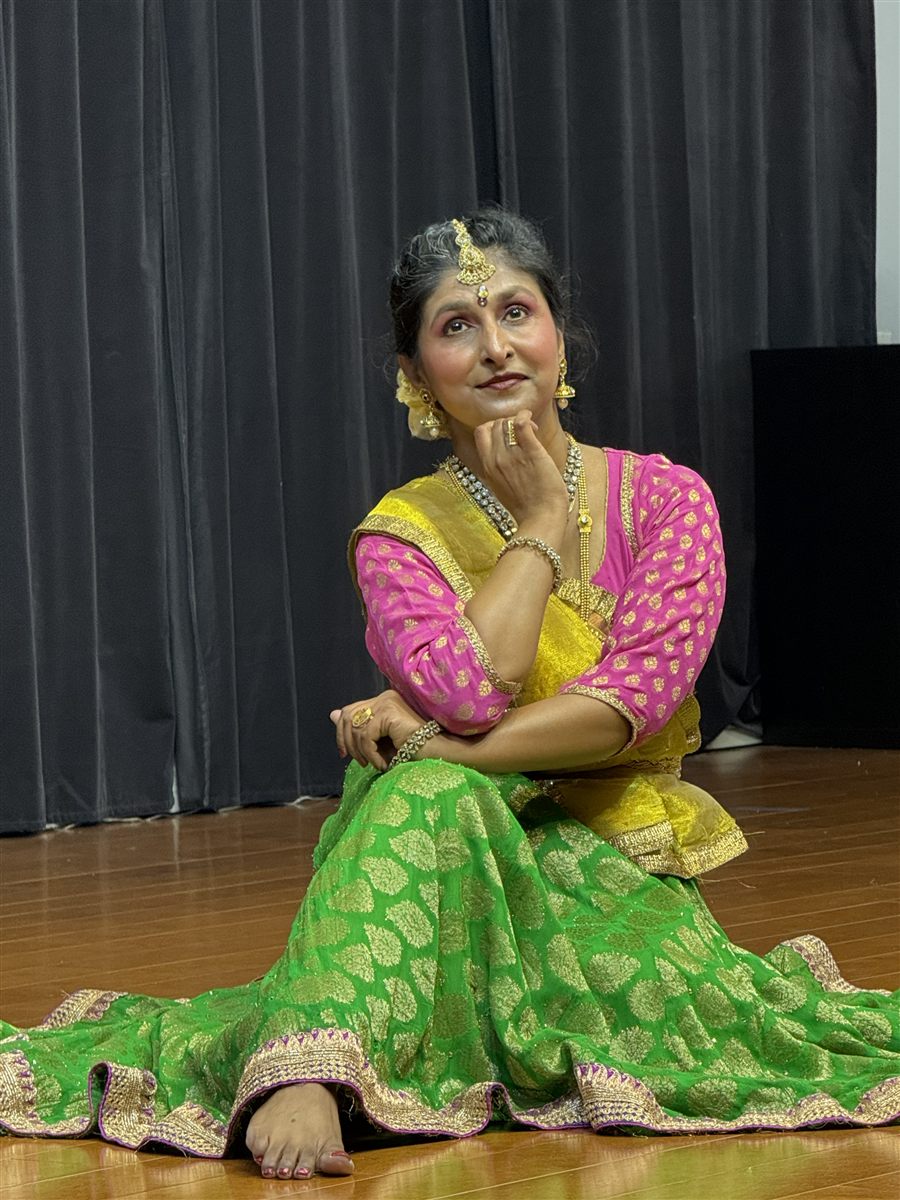 Sapna Adhikari in expressive Kathak abhinaya pose seated on stage in pink and green lehenga with gold jewelry